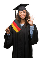 Young hispanic woman wearing graduation uniform holding flag of Germany doing ok sign with fingers,...