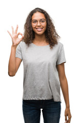 Beautiful young hispanic woman wearing glasses smiling positive doing ok sign with hand and...