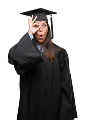 Young hispanic woman wearing graduated cap and uniform doing ok gesture shocked with surprised face, eye looking through fingers. Unbelieving expression.