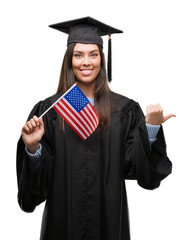 Young hispanic woman wearing graduated uniform holding flag of america pointing and showing with thumb up to the side with happy face smiling