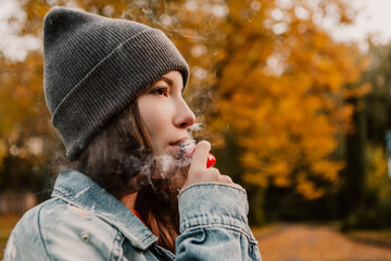 Young woman outside autumn park fall and smoking tobacco device electronic cigarette heater. Smoke...