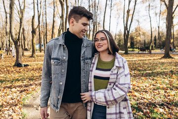 Heterosexual caucasian young loving couple walking outside in the city park in sunny weather, hugging smiling kissing laughing spending time together. Autumn, fall season, orange yellow red maple leav