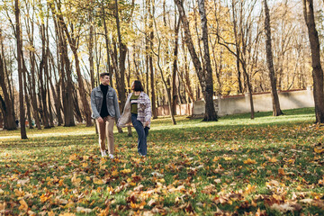 Fototapeta premium Heterosexual caucasian young loving couple walking outside in the city park in sunny weather, hugging smiling kissing laughing spending time together. Autumn, fall season, orange yellow red maple leav