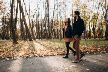 Fototapeta premium Heterosexual caucasian young loving couple walking outside in the city park in sunny weather, hugging smiling kissing laughing spending time together. Autumn, fall season, orange yellow red maple leav
