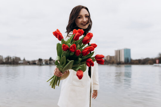 Attractive brunette woman walking on the beach shore in moody cloudy windy weather with bouquet of red tulips flowers, dressed in white suit jacket. International Women`s Day 8th March concept
