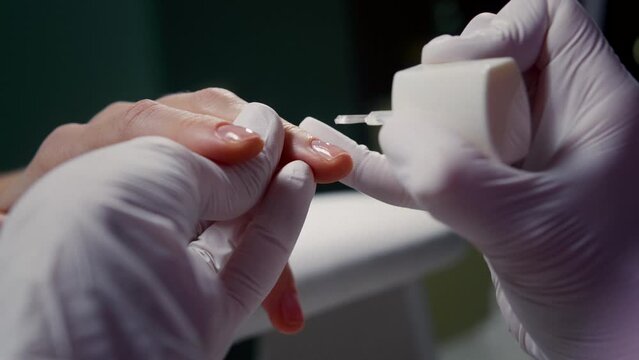 Master in gloves applies base polish on customer nail with brush in beauty salon closeup. Beautician uses clear laquer for woman fingernails in manicure shop. Cosmetologist works with client