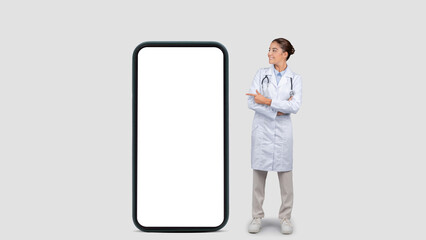 Happy caucasian millennial doctor in white coat point finger at big phone with empty screen
