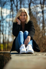 Woman in her mid-fifties with blonde long hair in a portrait, dressed in a black jacket, white T-shirt, blue jeans and white sneakers, outside in autumnal nature.