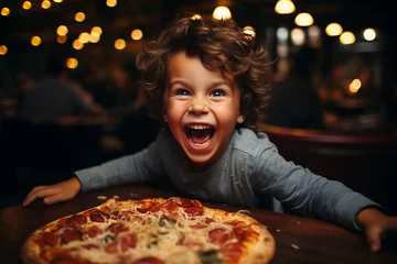 Foto op Aluminium  A happy laughing curly-haired boy at the table is eating fresh pizza, holding food in his hands. A joyful atmosphere in the pizzeria © Irina Afanaseva