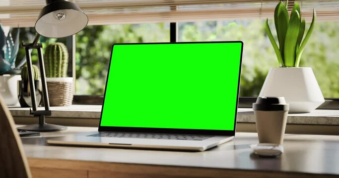 Laptop with a blank screen is placed on a stylish wooden desk within a loft-style interior, with green spaces in the background visible through the window. The camera moves smoothly from the bottom