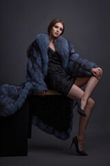 A young girl with exquisite makeup in a blue fur coat, a short black dress and black stylish shoes poses while sitting in the studio