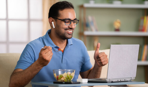 Happy indian man eating fruit salad by showing thumbs up on online video call at home - concept of satisfaction, communication and healthy eating. - Powered by Adobe