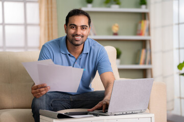 Happy Indian man checking financial documents or insurance bills looking at camera while working on laptop at home - concept of Budget tracking, Expense management and Contract review - obrazy, fototapety, plakaty