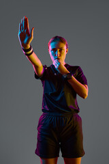 Young woman referee blowing whistle with serious expression against grey studio background in neon...