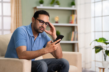 Shocked indian man got bad news while using mobile phone on sofa at home - concept of Financial distress, Loan cancellation and Uncertain future