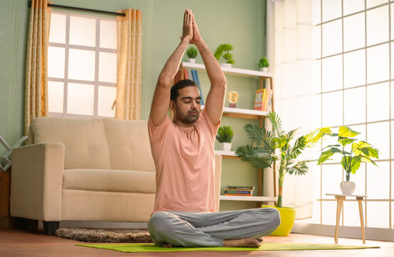 Indian man doing namaste pose yoga or exercise with eyes closed while sitting on floor at home - concept of morning routine, self care and health care