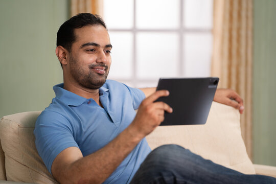 Happy indian young man watching digital tablet while sitting on sofa at home - concept of technology, relaxation and social media