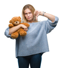 Young caucasian woman holding cute teddy bear over isolated background with angry face, negative sign showing dislike with thumbs down, rejection concept