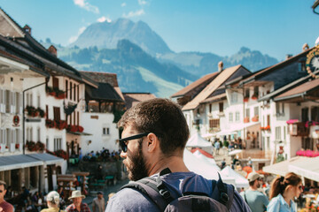 Young caucasian man tourist with beard and wearing sunglasses traveling with backpack exploring the european city Gruyères in Fribourg, Switzerland on summer season. SHOTLISTtravel. - obrazy, fototapety, plakaty