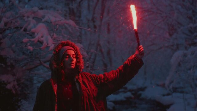 Portrait: A young girl holding a burning signal fire. A woman in a winter forest lights a red rescue flare. Snow on the trees, blue hour. Female person gives a distress signal. Snowy weather