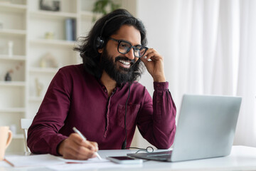 Obraz premium Young indian man wearing headset using laptop and smiling while taking notes