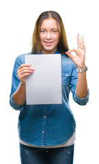 Young caucasian woman holding blank paper sheet over isolated background doing ok sign with fingers, excellent symbol