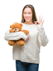 Young caucasian woman holding teddy bear over isolated background doing ok sign with fingers, excellent symbol