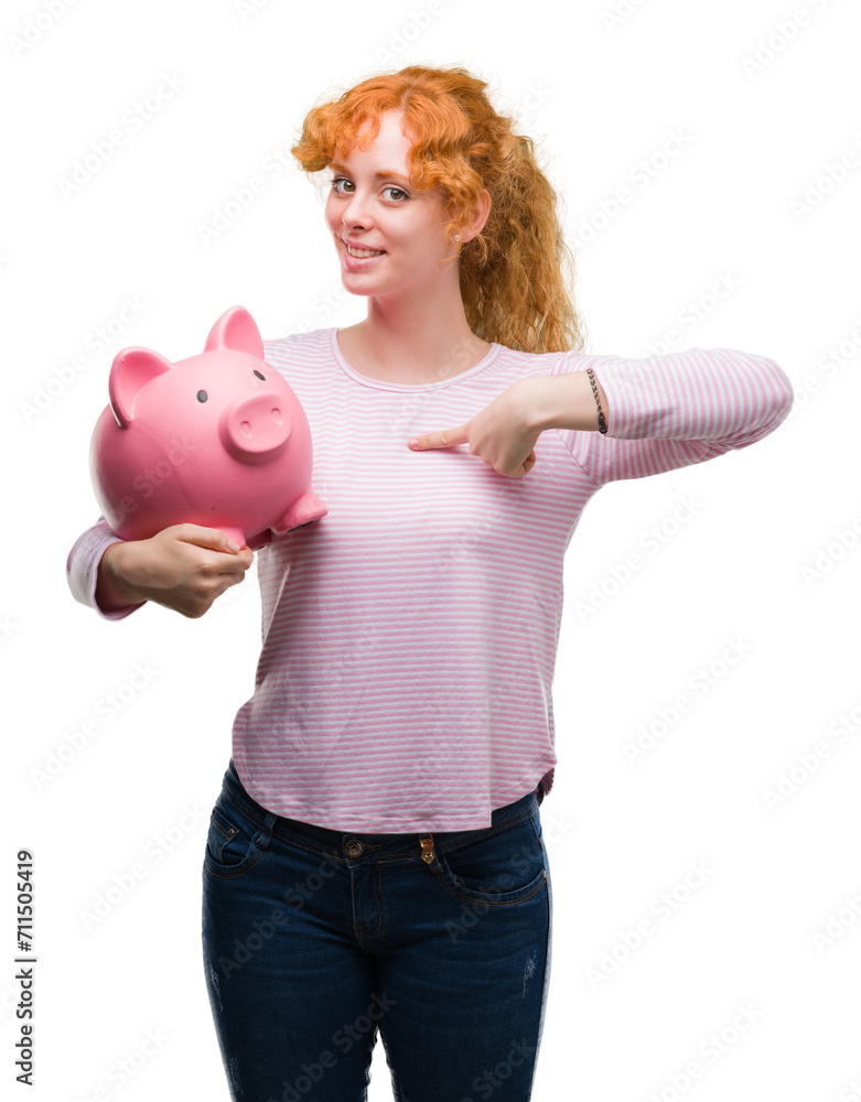 Sticker young redhead woman holding piggy bank with surprise face pointing finger to himself