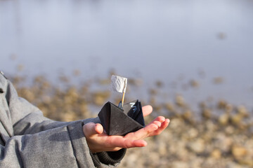A boy holds a paper black boat in his hands against the background of a stone beach in winter.