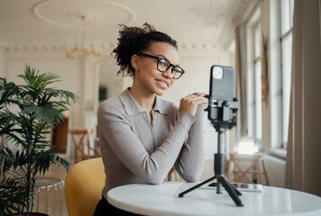 Smiling woman with glasses filming a blog on her smartphone in a bright, elegant room.