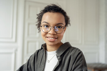 Confident young woman with glasses and curly hair smiling in a well-lit, classic room.