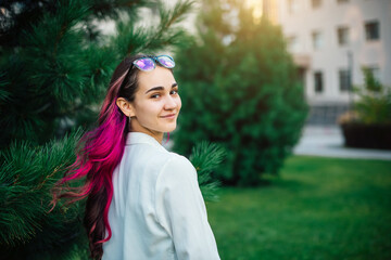 Naklejka premium A beautiful high school student with purple hair smiles and looks at the camera. Portrait of a girl with glasses in the park on the background of the university