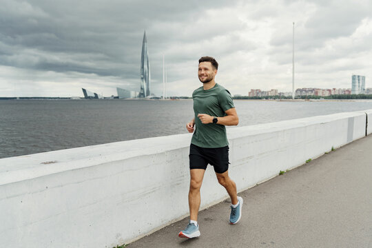 Smiling man in fitness attire running along a riverside promenade, with the dramatic cityscape and cloudy sky in the background. - Powered by Adobe