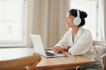 Relaxed woman in casual attire enjoying music with headphones while working on her laptop.