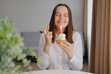 Hopeful smiling cheerful Caucasian brown haired woman celebrating birthday with cake and candle at home interior crossing her fingers making wish during her holiday celebration