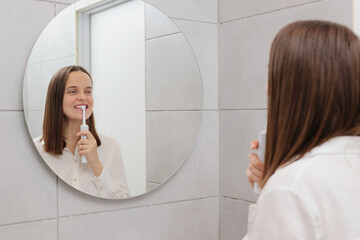 Clean and healthy mouth. Perfect oral hygiene. Dental protection methods. Morning oral care routine. Caucasian young adult woman brushing teeth in bathroom in front of mirror