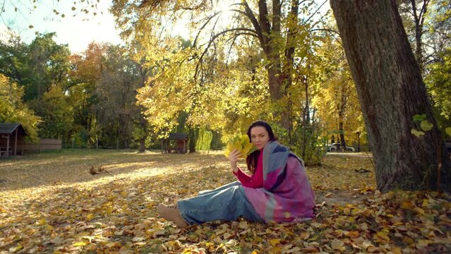 Happy smiling woman on picnic in autumn park. The girl makes a fool of herself and smiles, covering her face with a bouquet of puffed leaves. High quality 4k footage