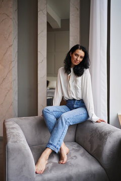 Capturing sophistication, the young woman in jeans and a white shirt sits on the grey chair's backrest by the window in a stylish interior, embodying modern elegance.