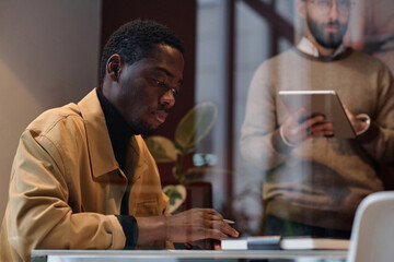 Black man in casual outfit working at desk in office, his caucasian colleague standing on background