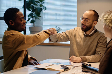 Joyful biracial male colleagues giving each other fist bump, their female coworker sitting nearby - Powered by Adobe