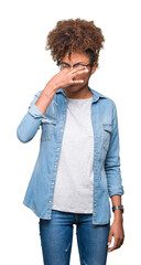 Beautiful young african american woman wearing glasses over isolated background smelling something...