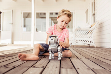 Young boy sitting on a deck playing with a robot dog. Cape Town, South Africa