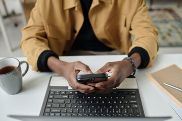 Hands of unrecognizable black man typing on smartphone while sitting in front of laptop at desk