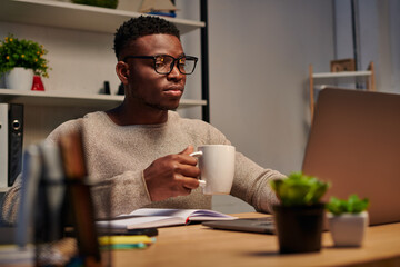 focused african american freelancer in eyeglasses holding coffee cup and working on laptop at home