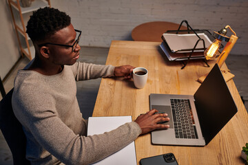 african american man in eyeglasses holding coffee cup and working on laptop in home office at night