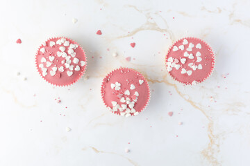 Pink glazed cupcakes decorated heart shape sprinkles on white golden marble background. Romantic sweet dessert, valentine muffins top view.