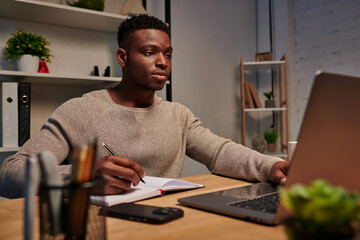 Fototapeta premium african american freelancer looking at laptop and writing in notebook working from home at night