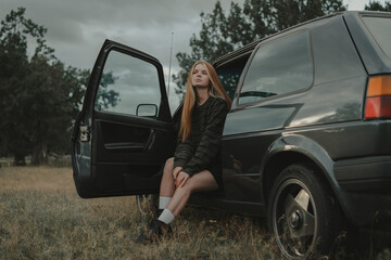 A redheaded girl in a green sweater sits in a retro car. The concept of free and cyclical fashion.