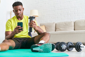 young african american man with sports bottle browsing internet on smartphone on fitness mat at home