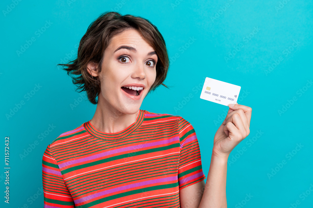 Sticker Photo of overjoyed woman with bob hairstyle dressed striped t-shirt holding debit card in fingers isolated on turquoise color background